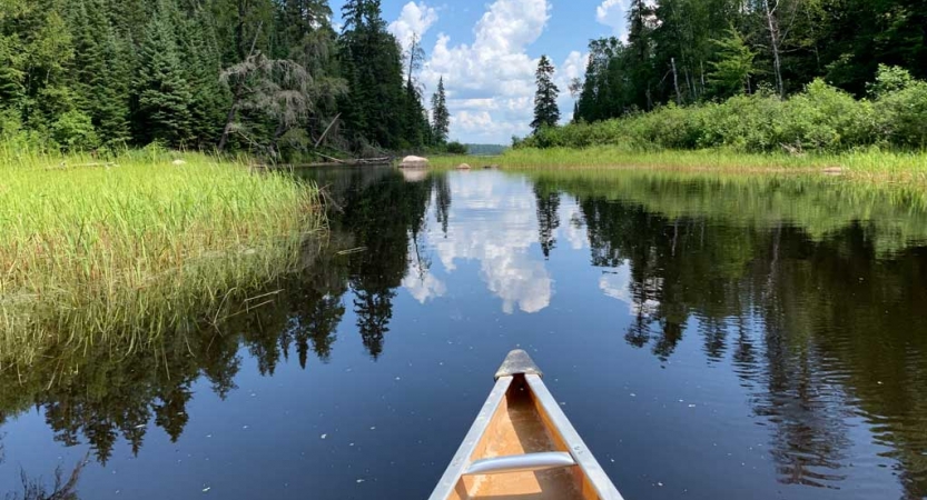 The tip of a canoe appears in the foreground, resting on calm still water that reflects the blue sky dotted with clouds. 